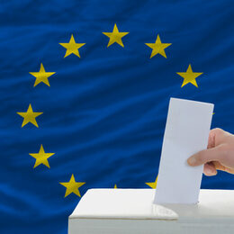 Das Bild zeigt im Hintergrund eine Hand, die ein Stück Papier in eine Wahlurne steckt. Im Hintergrund die Europaflagge.