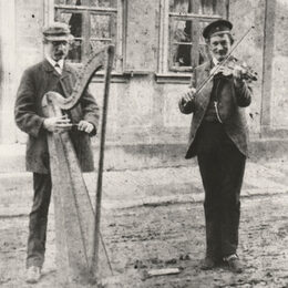 Das Bild aus dem 19. Jahrhundert zeigt zwei Klesmer-Musiker auf einer Straße vor einem Haus. Einer spielt Harfe, der andere Geige.